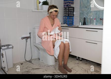 Eine Frau sitzt auf einer Toilette im Badezimmer. Sie trägt einen Bademantel und liest ein Blatt Papier. Verschiedene Badgegenstände sind um sie herum sichtbar. Stockfoto