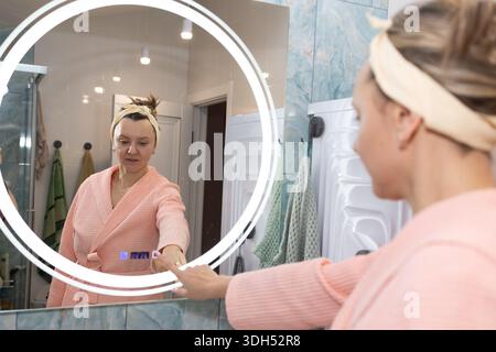 Eine Frau in einem Bademantel steht vor einem eleganten Spiegel in ihrem Badezimmer. Sie schaut auf den Bildschirm, während sie ihn berührt, und beginnt ihre Morgenroutine. Stockfoto
