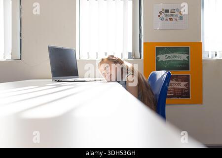 Junge Frau, die auf blauem Stuhl im Klassenzimmer sitzt, Kopf auf dem Tisch liegend, Laptop inaktiv, Kopierraum Stockfoto