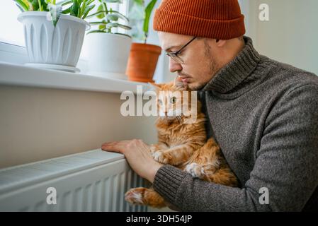 Ein Mann und eine Ingwerkatze wärmen sich mit einem Heizkörper auf, spüren die Kälte des Winters und hohe Heizrechnungen, verwenden Methoden wie das Tragen warmer Kleidung und Kuscheln, um Energiekosten zu sparen Stockfoto