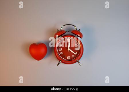 Rotes Herz und schwarzer Wecker auf blauer symbolischer Paarung, die auf die Verbindung der Liebe zu Zeit, Dringlichkeit oder dauerhafter emotionaler Präsenz hindeutet. Stockfoto