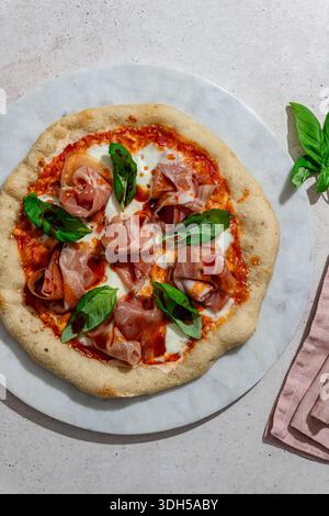 Blick von oben auf eine frisch gebackene Pizza mit Schinkenscheiben, frischen Basilikumblättern und cremigem Burrata-Käse, serviert auf einer Marmoroberfläche für einen Authenti Stockfoto