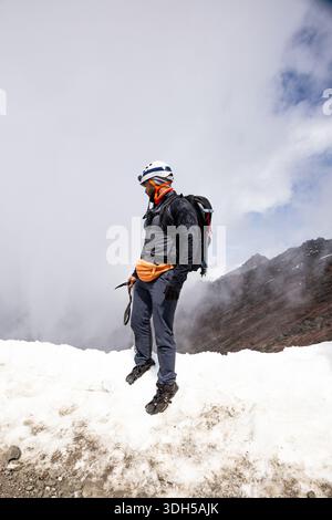 Ein Bergsteiger steht auf einem schneebedeckten Gipfel, ausgestattet mit unverzichtbarer Ausrüstung und einem Helm, und blickt in die nebelige Landschaft. Die Atmosphäre fängt den Nervenkitzel ein Stockfoto
