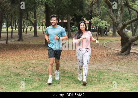 Ein Paar joggt gemeinsam durch einen üppigen Park in Lateinamerika und genießt Outdoor-Übungen inmitten hoher Bäume und eines malerischen Weges, der eine Heilung verkörpert Stockfoto