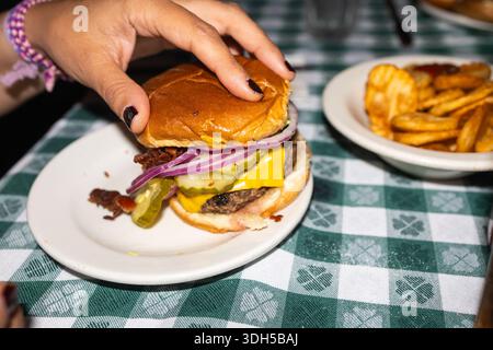 Abgeschnittene, nicht erkennbare weibliche Hand, die einen saftigen Cheeseburger mit Gurken und Zwiebeln auf einem weißen Teller hält. Ein appetitlicher Teller mit lockigen Pommes frites ist daneben Stockfoto