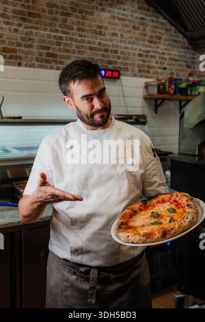 Ein erfahrener Pizzamacher in einer Pizzeria präsentiert stolz eine köstliche neapolitanische Pizza, frisch aus dem Ofen. Er trägt ein knuspriges weißes Koch-Outfit, symbolizin Stockfoto