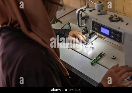 Eine asiatisch-muslimische Schneiderin arbeitet geschickt an einem handgefertigten Stoffdesign mit einer Nähmaschine in ihrer gemütlichen Heimwerkstatt. Kreativität und Tradition Stockfoto