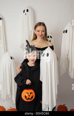 Eine Frau und ein Junge genießen Halloween-Festlichkeiten, umgeben von verspielten Geisterdekorationen und Kürbissen. Der Junge hält einen kürbisförmigen Eimer, verkörpert Stockfoto