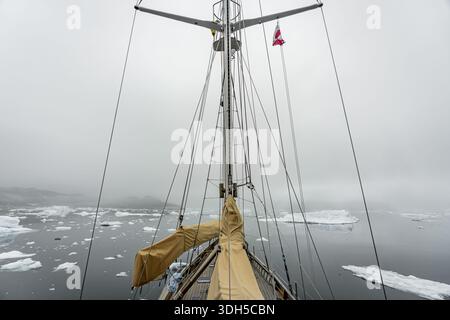 Ein Segelboot navigiert durch das eisige Wasser in Grönland, umgeben von treibenden Eisbergen unter einem bewölkten Himmel. Die ruhige arktische Landschaft sorgt für eine kühle Atmosphäre Stockfoto