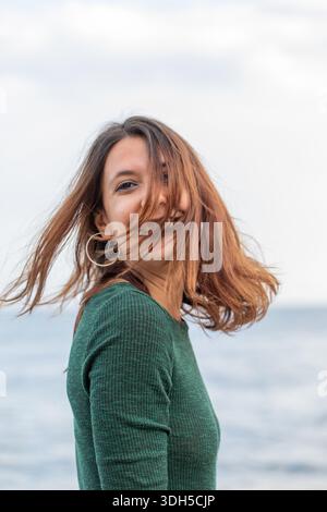 Eine fröhliche Frau mit fließenden Haaren, die einen grünen Pullover trägt, lächelt, während sie einen luftigen Tag am Meer genießt. Das ruhige Meer dient als beruhigender Rücken Stockfoto