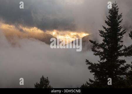 Ein atemberaubender Blick auf nebelbedeckte Berge bei Sonnenaufgang. Das goldene Licht durchdringt dicke Wolken und erzeugt einen dramatischen Kontrast bei dichtem Nebel Stockfoto