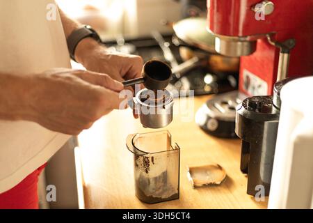 Zugeschnittener, nicht erkennbarer Mann, der Kaffee mit einem Portafilter auf einer Holztheke in einer Küche zubereitete, mit einer roten Kaffeemaschine für die Zubereitung von frischem Espresso Stockfoto