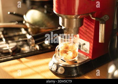 Frischer Kaffee brüht sich in einer roten Kaffeemaschine, füllt eine Tasse aus klarem Glas mit reichhaltigem, dampfendem Espresso, während das Sonnenlicht das gemütliche Kit sanft beleuchtet Stockfoto