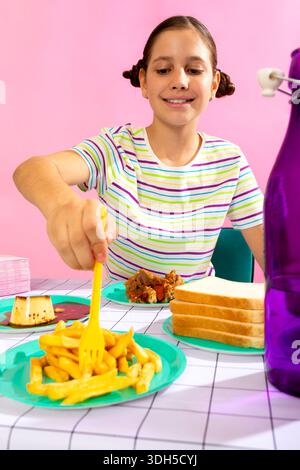 Ein fröhliches Mädchen sitzt an einem Tisch mit einer Vielzahl von Speisen, darunter Pommes und Desserts, vor einem farbenfrohen Hintergrund. Ihr Lächeln spiegelt ihre Freude wider Stockfoto