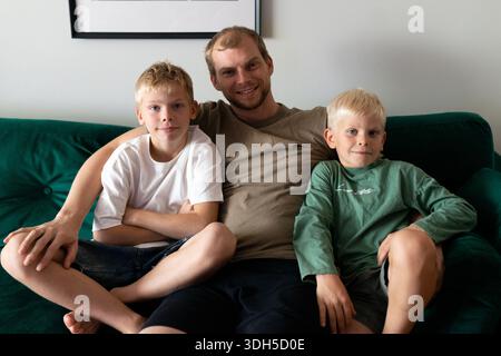 Eine fröhliche Familie mit drei Personen genießt eine gute gemeinsame Zeit auf einer komfortablen grünen Couch zu Hause. Der Vater und die Söhne teilen ein Lächeln, drücken Wärme aus und Stockfoto