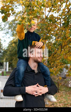 Ein fröhlicher Vater trägt seinen glücklichen Sohn auf seinen Schultern, während er durch einen lebendigen Park schlendert, umgeben von der Schönheit der Natur, und einen Moment der fami festnimmt Stockfoto