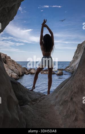 Eine Frau genießt den ruhigen Blick auf den italienischen Strand, steht auf einer felsigen Felsformation mit erhobenen Armen, unter einem klaren blauen Himmel und dem ruhigen Meer in Th Stockfoto