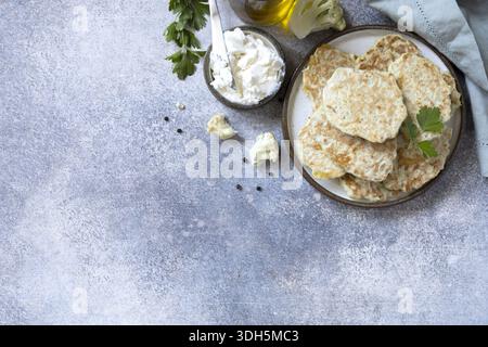 Blumenkohlpfannkuchen garniert mit frischen Kräutern und serviert mit Sauerrahm. Präsentiert auf einer rustikalen Oberfläche, die Szene umfasst ein weiches Tuch, Stockfoto