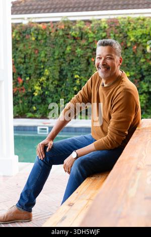 Reifer Mann sitzt auf Holzbank am Poolrand mit braunem Pullover, Schuhen und Armbanduhr Stockfoto