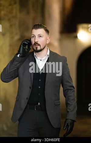 Ein scharf gekleideter Mann in einem maßgeschneiderten Anzug unterhält sich auf seinem Telefon, umgeben von dem Charme der antiken Architektur, die nachts von sanften Lichtern beleuchtet wird. Stockfoto