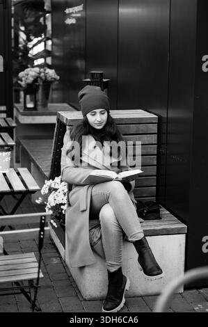 Unter einem sanften Sonnenuntergang sitzt eine junge Frau bequem auf einer Bank, vertieft in ihr Buch, umgeben von blühenden Blumen und friedlicher Stadtansprache. Stockfoto