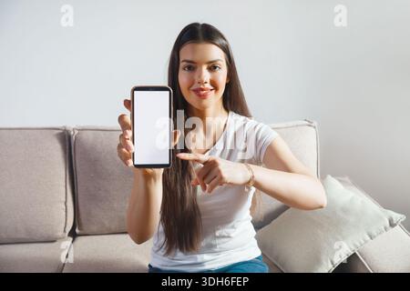 Eine Frau sitzt auf einer Couch und zeigt auf ein Smartphone, das sie hält. Sie blickt mit einem Lächeln direkt in die Kamera. Die Inneneinrichtung hat einfache Möbel Stockfoto