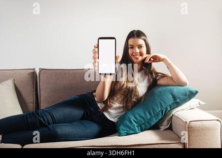 Eine Frau sitzt auf einer Couch, lächelt und hält ein Smartphone mit einem leeren Bildschirm. Sie hat lange Haare und trägt legere Kleidung, was ein entspanntes AT schafft Stockfoto