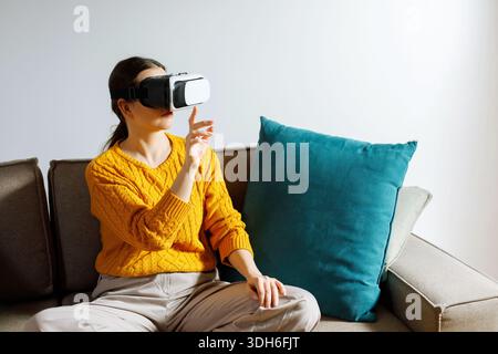 Eine Frau sitzt leise auf einer Couch, während sie ein Virtual-Reality-Headset trägt. Sie scheint konzentriert und mit der virtuellen Erfahrung in ihrem Wohnraum beschäftigt zu sein Stockfoto