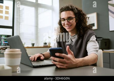 Junger Erwachsener kaukasischer Mann lächelt, während er drinnen Smartphone und Laptop benutzt Stockfoto