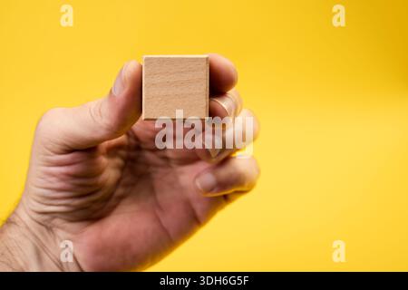 Eine Hand, die einen Holzblock vor einem gelben Hintergrund hält. Zeigt den Holzblock zur Kamera. Konzept des unternehmerischen und unternehmerischen Erfolgs. Stockfoto