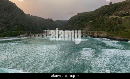 Aus der Vogelperspektive auf die alte Eisenbahnbrücke über die Mündung des Kaaimans River, wo die türkisfarbene Umarmung des Ozeans auf die zerklüftete Küste, Wilderness, Weste, trifft Stockfoto