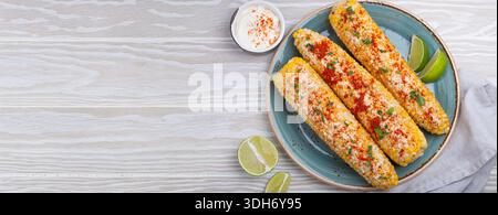 Drei gegrillte Maiskolben mit Gewürzen, Mayonnaise und Cotija werden auf einem Teller mit Limettenscheiben und Chili-Pulver-Sauerrahm serviert Stockfoto