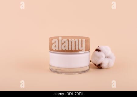 Ein Glas mit Feuchtigkeitscreme und Baumwollzweig auf beigefarbenem Hintergrund. Naturschönheitsprodukt, Bio-Kosmetik kreatives Konzept. Detaillierter Behälter aus weißem Glas Stockfoto