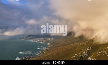 Aus der Vogelperspektive sehen Sie dramatische Wolken, die sich an der zerklüfteten Küste in der Nähe von Camps Bay, wo das türkisfarbene Meer auf die goldenen Hügel trifft, Kapstadt, Western C Stockfoto