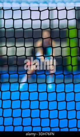 Nahaufnahme eines schwarzen Padelnetzes im Fokus mit einem unscharfen Spieler, der auf einem blauen Spielfeld auf einen Ball im Hintergrund wartet. Stockfoto