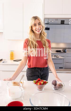 Frau in rotem Hemd, die hinter der Marmorinsel in der Küche steht und Erdbeeren und Milch zubereitete Stockfoto