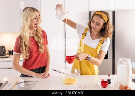 Freundinnen in rotem T-Stück, gelbem Overall, der Milch aus der Kanne in die rote Tasse in der Küche gießt Stockfoto