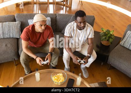 Verschiedene männliche Freunde sitzen zu Hause auf grauem Sofa und spielen Spiele, halten Controller, teilen Chips Stockfoto