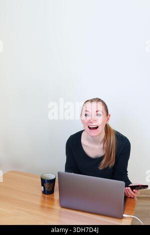 Aufgeregte Frau, die mit Laptop und Smartphone an einem Schreibtisch sitzt. Positive Überraschung, Online-Erfolg und modernes Home-Office-Konzept. Stockfoto