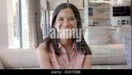 Lächelnde Frau mit ärmellosem pinkem Oberteil, die zu Hause auf einer beigen Couch mit blaublauem Code-Overlay sitzt Stockfoto
