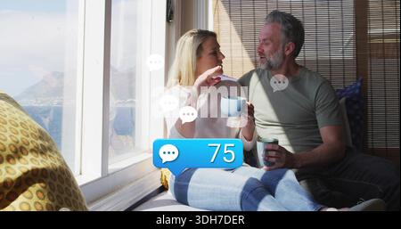 Sitzendes Paar, das Tassen hält und zu Hause auf dem Fenstersitz plaudert, in lässiger Kleidung, Abzeichen 75 Stockfoto