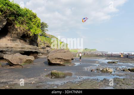 Tanah Lot, Bali – 08. August 2025: Farbenfroher Drachen schwingt über die felsige Küste, während Besucher bei Ebbe unter verstreuten Wolken spazieren gehen. Stockfoto