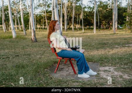 Seitenansicht einer rothaarigen Frau, die auf einer roten Bank in einem Park sitzt, ruhig und nachdenklich. Stockfoto