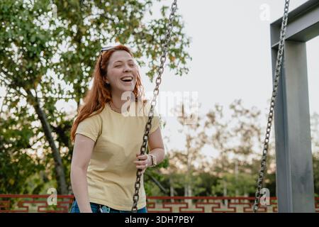 Lachende junge rothaarige Frau mit Schaukelketten auf einem Spielplatz in einem Park, fröhlicher Outdoor-Lifestyle. Stockfoto