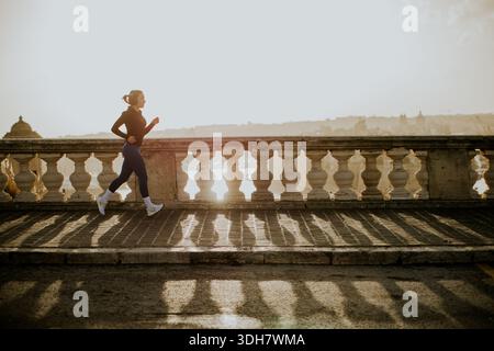 Eine Frau joggt während des Sonnenuntergangs auf einem Fußweg am Meer in Malta. Das Sonnenlicht leuchtet auf und erzeugt lange Schatten und eine warme Atmosphäre um sie herum Stockfoto