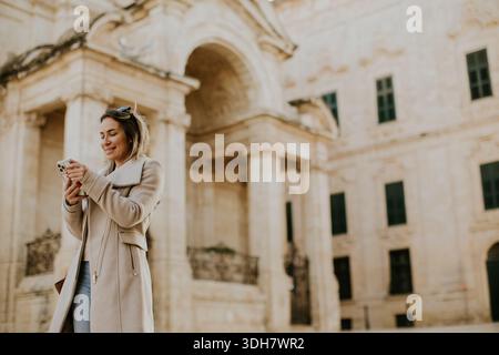 Eine Frau läuft durch ein historisches Viertel in Malta, während sie auf ihr Handy schaut. Sie steht vor großen Steinhäusern und genießt ihre Zeit im Freien Stockfoto