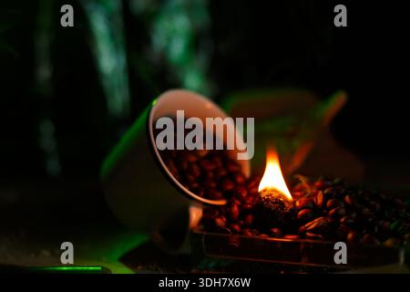 Kaffeebohnen, die aus der Tasse neben einem kleinen brennenden Emblem verschüttet werden, erzeugen eine aromatische Szene Stockfoto