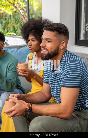 Verschiedene Freunde sitzen auf dem Sofa und halten Tassen am Fenster, bärtiger Indianer mit gestreiftem Hemd Stockfoto