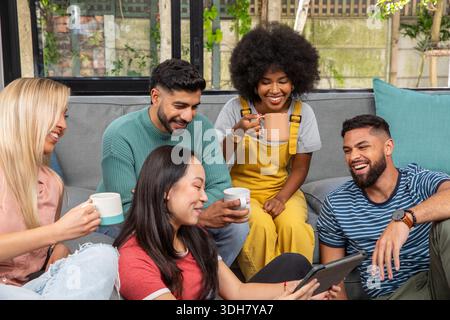 Verschiedene Freunde sitzen zu Hause auf grauem Sofa und Boden, lehnen sich hinein, teilen sich ein Tablet, halten Tassen in der Hand Stockfoto