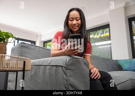Chinesin sitzt auf grauem Sofa im Wohnzimmer in rotem T-Shirt und hält ein schwarzes Smartphone und lächelt Stockfoto